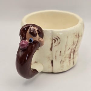 May include: A ceramic mug with a cream-colored body featuring brown textured markings resembling tree bark. The handle is shaped like a brown figure with a small purple nose and a single visible blue eye, extending from the side of the mug.
