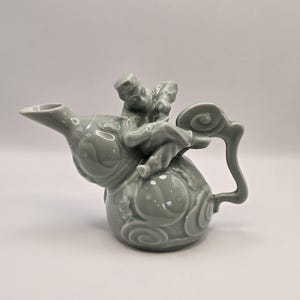 May include: A celadon-colored ceramic teapot with a whimsical design. The teapot features a sculpted figure riding atop the body, with a curved handle and spout. The body of the teapot is decorated with swirling patterns.