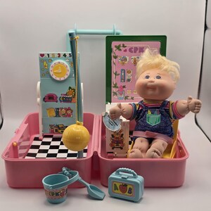 May include: A Cabbage Patch Kids doll with blonde hair wearing denim overalls, seated in a pink plastic playset case. The set includes a clock panel, a yellow ball on a pole, a blue bucket with a shovel, and a small blue lunchbox with an apple icon.