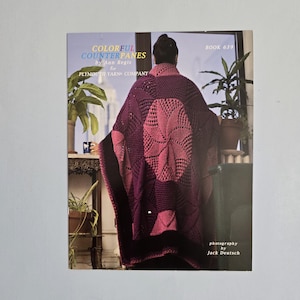 May include: A book cover featuring a woman wearing a large, crocheted shawl in shades of purple and pink. The book title reads "COLORFUL COUNTERPANES" by Ann Regis for PLYMOUTH YARN COMPANY. Photography by Jack Deutsch.