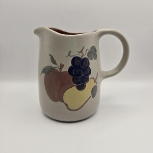 May include: A beige ceramic pitcher featuring a painted design of a brown apple, a yellow pear, and a cluster of dark purple grapes with green leaves. The pitcher has a curved handle on the right side and a small pouring spout on the top rim.