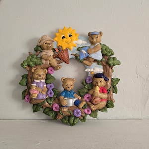 May include: A decorative wreath featuring five teddy bears in various outfits, surrounded by green leaves, purple flowers, and a smiling yellow sun. The bears are engaged in different activities, such as fishing and gardening. The wreath is made of a durable material.