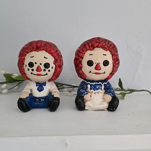 May include: A pair of vintage rag doll salt and pepper shakers. The dolls are sitting down and have red hair, blue eyes, and painted smiles. The boy doll is wearing a blue shirt and pants, while the girl doll is wearing a blue dress with white stars.