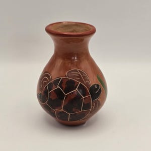 May include: A small, rounded terracotta vase with a smooth, polished finish. The vase features a hand-painted dark brown turtle design on its front, accented with fine white etched lines and a single green leaf detail near the turtle's shell.