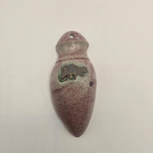 May include: A teardrop-shaped ceramic wall pocket with a speckled mauve and white glaze. The piece features a small hole at the top for hanging and a central embossed relief of a buffalo in a dark, metallic-toned glaze with teal accents.