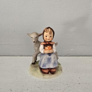 May include: A ceramic figurine of a young girl in a blue dress and brown shoes, holding a basket of flowers. A gray lamb stands beside her. The figurine is set on a grassy base.