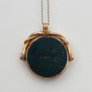 May include: A gold-coloured pendant necklace featuring a dark green stone with red flecks. The circular pendant has a twisted rope design around the stone. The chain is a delicate silver colour.