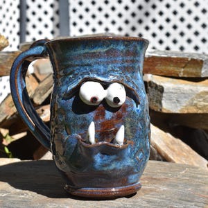 May include: A handmade ceramic mug with a glossy blue and brown glaze, featuring a sculpted monster face with two protruding white eyes, heavy eyelids, and two sharp white fangs extending from the mouth area, complete with a curved handle.