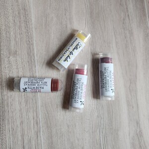 Puede incluir: Cuatro tubos de b&aacute;lsamo labial con tapas transparentes est&aacute;n dispuestos sobre una superficie de madera. Los tubos est&aacute;n etiquetados con las palabras "Red", "Rose" y "Tallow lip balm". Los b&aacute;lsamos labiales son de varios tonos de rojo y amarillo.