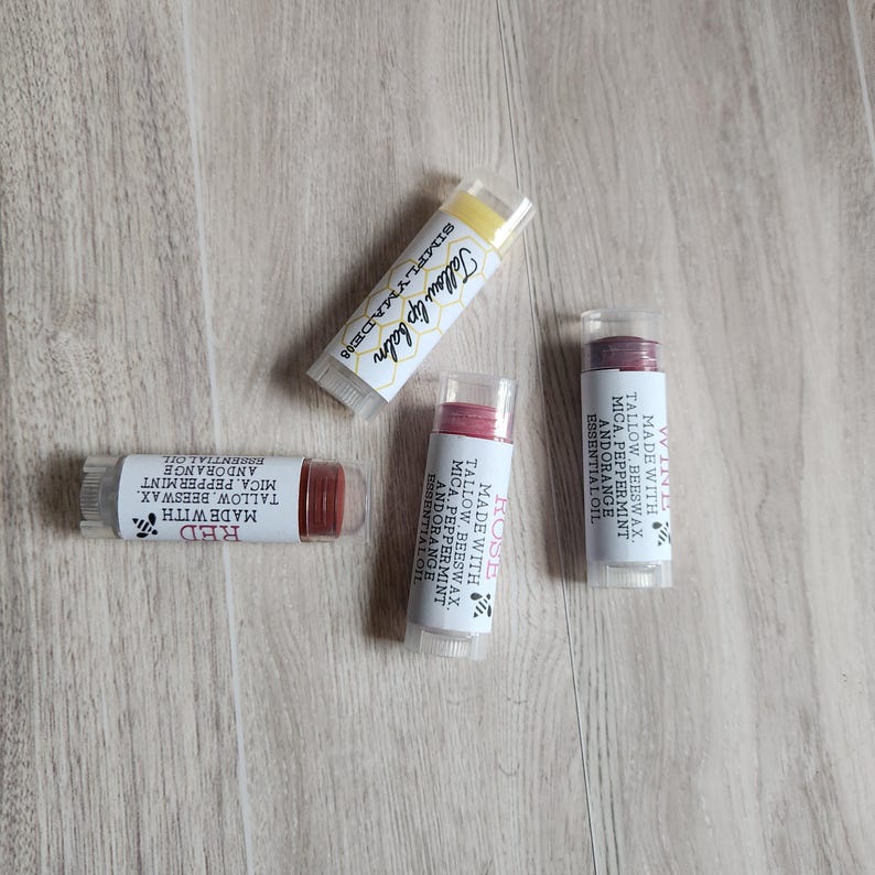 Puede incluir: Cuatro tubos de b&aacute;lsamo labial con tapas transparentes est&aacute;n dispuestos sobre una superficie de madera. Los tubos est&aacute;n etiquetados con las palabras "Red", "Rose" y "Tallow lip balm". Los b&aacute;lsamos labiales son de varios tonos de rojo y amarillo.