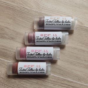 May include: Four tinted tallow lip balms with SPF 15. Each balm is in a clear tube with a white label that reads "SPF 15 Tinted Tallow lip balm SIMPLYMADE08".