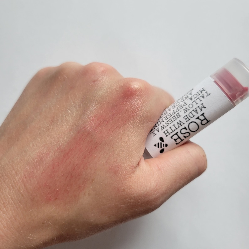 Puede incluir: Un tubo transparente de b&aacute;lsamo labial con una etiqueta blanca que dice "ROSE MADE WITH TALLOW, BEESWAX, MICA, PEPPERMINT, AND ORANGE". El b&aacute;lsamo es de color rosa claro, y una muestra del b&aacute;lsamo es visible en la mano de la persona.