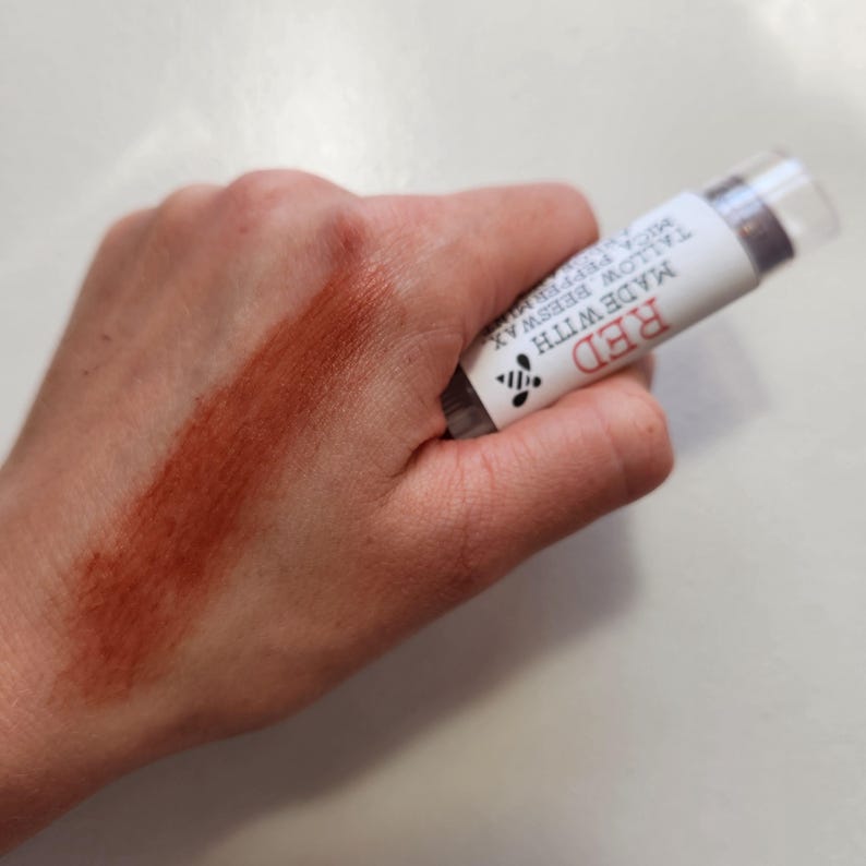 Puede incluir: Un labial rojo con tapa transparente se sostiene en una mano, con una muestra de color rojo en la piel. La etiqueta dice "RED MADE WITH TALLOW BEESWAX MICA PEPPERMINT". Un peque&ntilde;o gr&aacute;fico de abeja tambi&eacute;n est&aacute; en la etiqueta.
