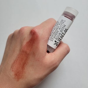 Puede incluir: Un tubo de b&aacute;lsamo labial transparente con una etiqueta blanca que dice "WINE" e incluye ingredientes. Una muestra del b&aacute;lsamo de color marr&oacute;n rojizo es visible en la mano. El b&aacute;lsamo contiene cera de abejas, sebo, mica, menta y aceites esenciales de naranja.