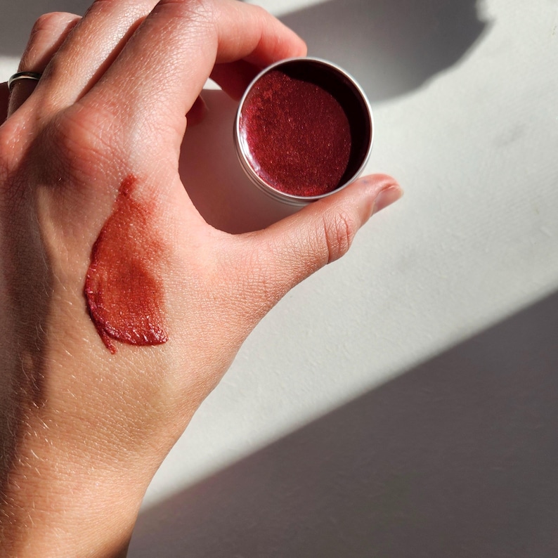 A metal tin of deep reddish lip balm or cream blush. The product has a shiny, glossy finish.  A swatch of the product is shown on a hand, demonstrating its color and texture. The balm appears to be richly pigmented and smooth.