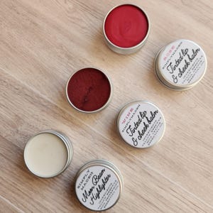 May include: Four small metal tins with lids containing different colored lip and cheek balms. The tins are labeled with the product names "Moon Beam Highlighter", "Wine", "Rose", and "Tinted Lip & Cheek Balm".