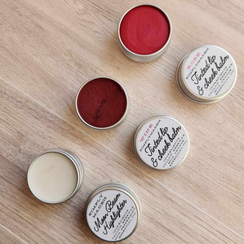 May include: Four small metal tins with lids containing different colored lip and cheek balms. The tins are labeled with the product names "Moon Beam Highlighter", "Wine", "Rose", and "Tinted Lip & Cheek Balm".