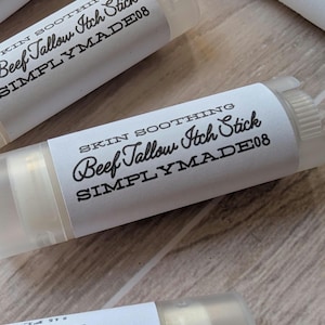May include: Several clear tubes of "Beef Tallow Itch Stick" are displayed, each with a white label. The labels have black text, including "Skin Soothing" and "SIMPLYMADE08". The tubes are arranged on a light-coloured wooden surface.
