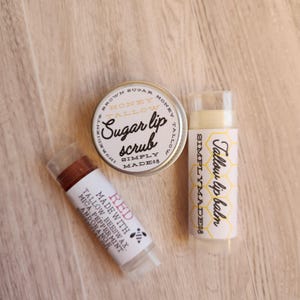 May include: Three lip care products: a small tin of honey and tallow sugar lip scrub, a clear tube of red lip balm, and a clear tube of tallow lip balm. The labels on the products say "Simply Made 08" and "Tallow Lip Balm".
