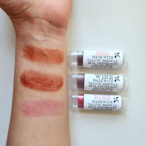 Three tinted lip balms in shades of red, wine, and rose. Each balm is in a clear tube with a white label indicating the color and ingredients: tallow, beeswax, mica, peppermint, and orange essential oil.  The labels also include the brand's logo, a small bee illustration.