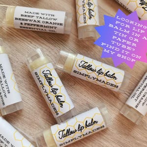 Puede incluir: Varios tubos de b&aacute;lsamo labial con la etiqueta "Tallow lip balm SIMPLYMADE08". Los tubos transparentes contienen un b&aacute;lsamo amarillo p&aacute;lido. Ingredientes: sebo de res, cera de abejas, naranja y aceite esencial de menta. El texto "Looking for lip balm in a tin or paper tube? Find it on my shop."