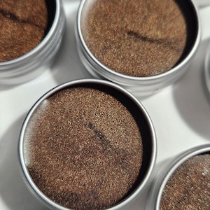 May include: Six small round metal tins filled with a brown shimmery substance. The tins have silver lids.