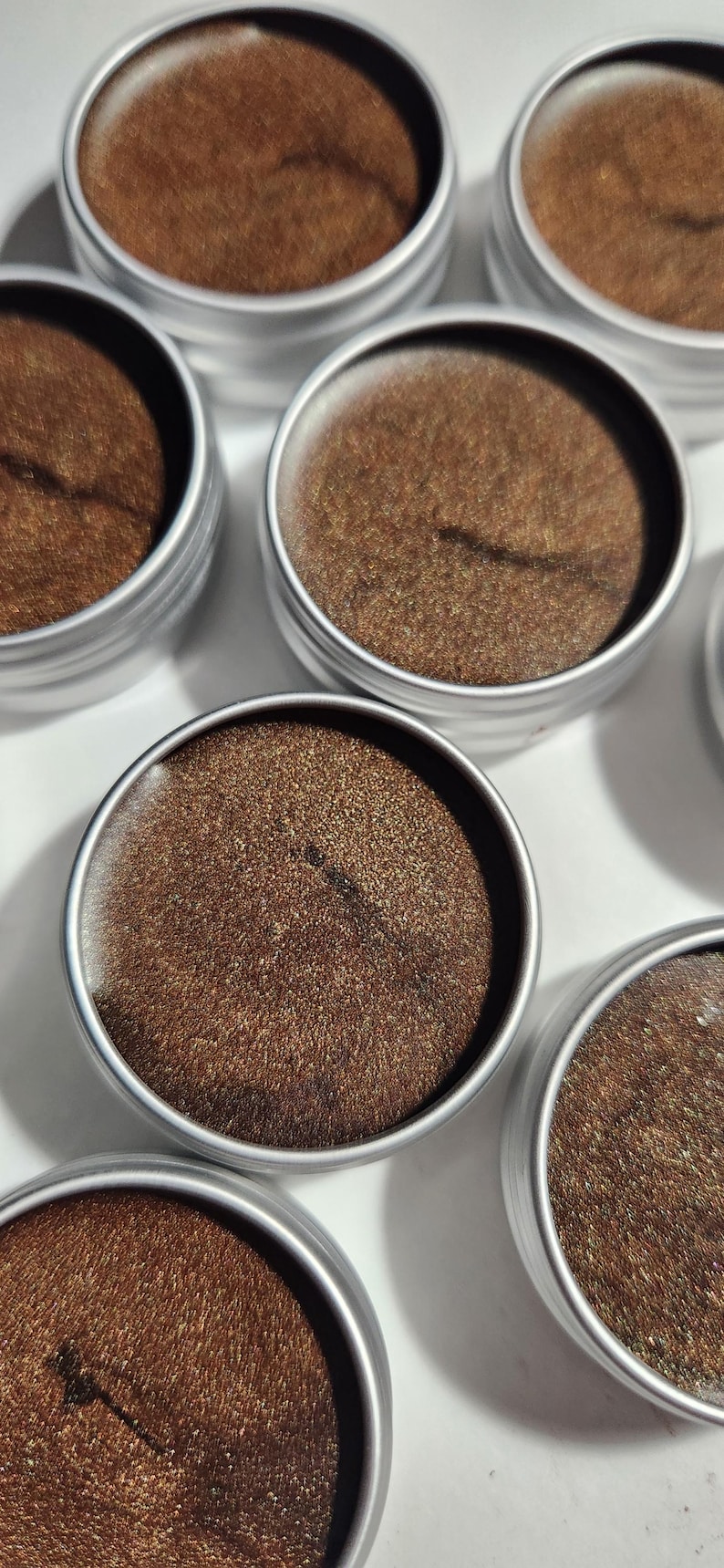 May include: Six small round metal tins filled with a brown shimmery substance. The tins have silver lids.