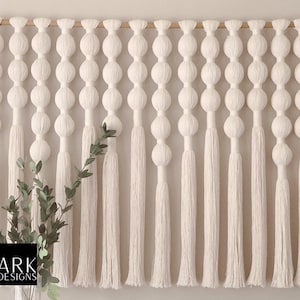K&ouml;nnte beinhalten: Wei&szlig;er Makramee-Wandbehang mit einer Holzstange oben. Der Wandbehang hat eine Reihe von wei&szlig;en Garnpompons und Quasten.