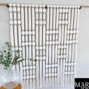 Könnte beinhalten: Weißer Makramee-Wandbehang mit einem geometrischen Muster aus weißen Perlen und Quasten. Der Wandbehang ist aus Baumwollgarn gefertigt und etwa 76 cm breit und 102 cm lang.