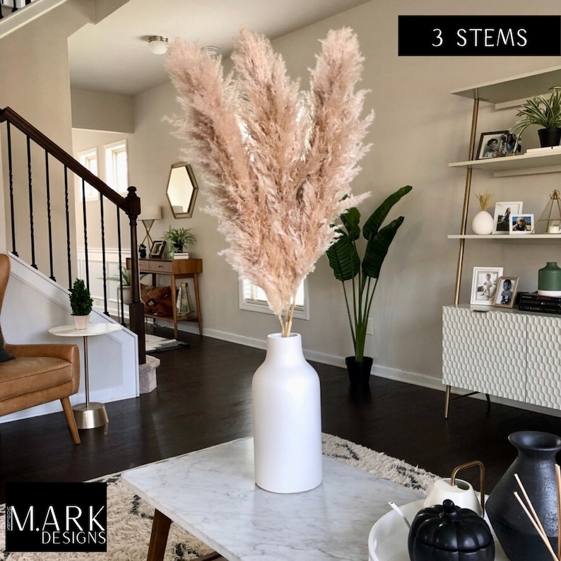 May include: Three stems of dried pampas grass in a white ceramic vase on a marble coffee table. The vase is tall and cylindrical with a rounded base. The pampas grass is a light beige color and is arranged in a loose, natural style. The text "3 STEMS" is visible in the upper right corner of the image.