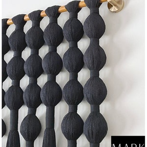 Op de afbeelding: Zwarte macram&eacute; wandhanger met een reeks grote, afgeronde knopen die aan een houten roede zijn opgehangen. De knopen zijn gelijkmatig verdeeld en verbonden door verticale strengen, waardoor een gestructureerd, decoratief element ontstaat. Een messingkleurige haak bevestigt de roede aan de muur.