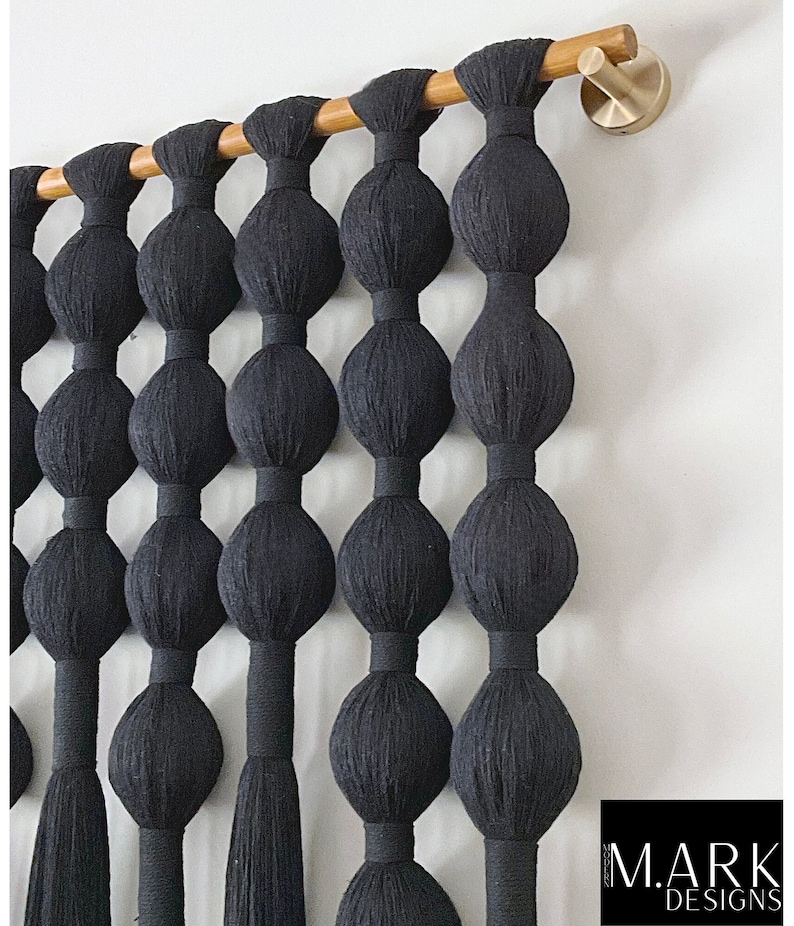 Op de afbeelding: Zwarte macram&eacute; wandhanger met een reeks grote, afgeronde knopen die aan een houten roede zijn opgehangen. De knopen zijn gelijkmatig verdeeld en verbonden door verticale strengen, waardoor een gestructureerd, decoratief element ontstaat. Een messingkleurige haak bevestigt de roede aan de muur.