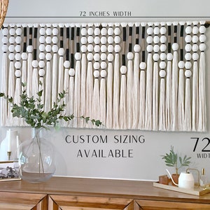 K&ouml;nnte beinhalten: Ein wei&szlig;er, schwarzer und beigefarbener Makramee-Wandbehang mit Pompons und Quasten. Der Behang misst 72 Zoll in der Breite und 36 Zoll in der H&ouml;he. Der Text "CUSTOM SIZING AVAILABLE" und "72X36" ist unter dem Behang gedruckt.
