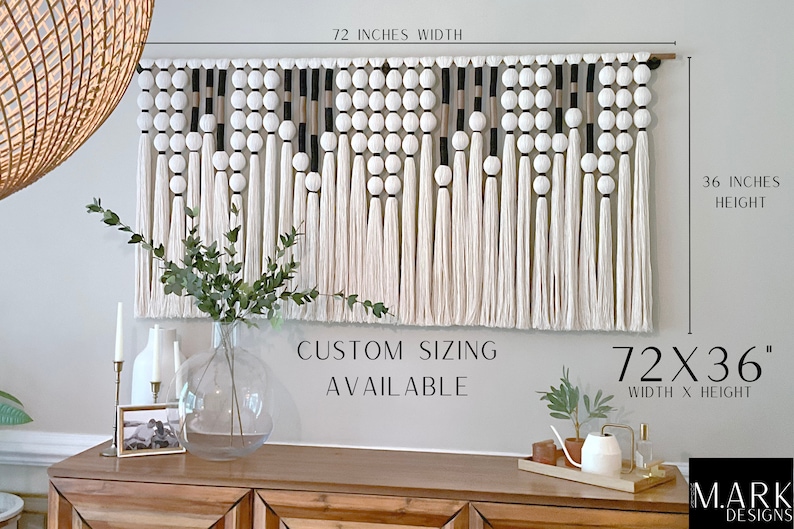 K&ouml;nnte beinhalten: Ein wei&szlig;er, schwarzer und beigefarbener Makramee-Wandbehang mit Pompons und Quasten. Der Behang misst 72 Zoll in der Breite und 36 Zoll in der H&ouml;he. Der Text "CUSTOM SIZING AVAILABLE" und "72X36" ist unter dem Behang gedruckt.