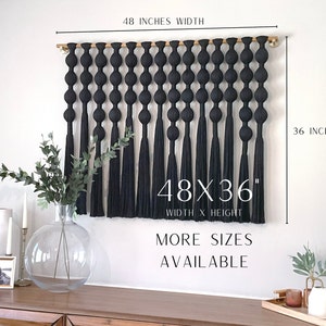 Op de afbeelding: Een zwarte macram&eacute; wandhanger, 122 cm breed en 91 cm hoog, met een reeks geknoopte elementen en lange kwasten. De tekst "48X36" en "MORE SIZES AVAILABLE" zijn zichtbaar. De wandhanger wordt op een witte muur getoond.