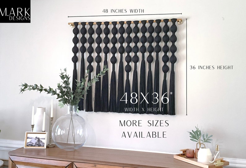 Op de afbeelding: Een zwarte macram&eacute; wandhanger, 122 cm breed en 91 cm hoog, met een reeks geknoopte elementen en lange kwasten. De tekst "48X36" en "MORE SIZES AVAILABLE" zijn zichtbaar. De wandhanger wordt op een witte muur getoond.
