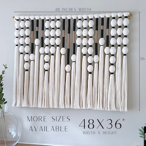 K&ouml;nnte beinhalten: Ein wei&szlig;er, schwarzer und beiger Makramee-Wandbehang mit Holzstab und wei&szlig;en Pompons. Der Wandbehang misst 122 cm in der Breite und 91 cm in der H&ouml;he. Der Text "MORE SIZES AVAILABLE" und "48X36" WIDTH X HEIGHT ist unter dem Wandbehang gedruckt. Der Text "MARK DESIGNS" ist in der unteren rechten Ecke gedruckt.