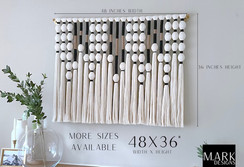 K&ouml;nnte beinhalten: Ein wei&szlig;er, schwarzer und beiger Makramee-Wandbehang mit Holzstab und wei&szlig;en Pompons. Der Wandbehang misst 122 cm in der Breite und 91 cm in der H&ouml;he. Der Text "MORE SIZES AVAILABLE" und "48X36" WIDTH X HEIGHT ist unter dem Wandbehang gedruckt. Der Text "MARK DESIGNS" ist in der unteren rechten Ecke gedruckt.