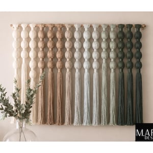 May include: A wall hanging featuring a series of vertical fiber strands suspended from a horizontal rod. Each strand consists of stacked, rounded fabric spheres transitioning in color from cream to tan, gray, and dark green, ending in long tassels.