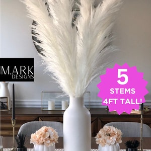 May include: Five white pampas grass stems in a white ceramic vase, 4 feet tall. The vase is on a wooden table with two small white pumpkins with peach colored flowers on top. The text "5 STEMS 4FT TALL!" is on a pink starburst shape. The text "WHITE" is on the table.