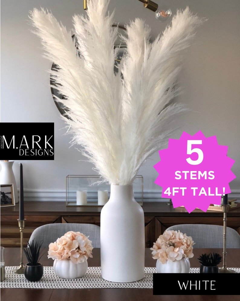 May include: Five white pampas grass stems in a white ceramic vase, 4 feet tall. The vase is on a wooden table with two small white pumpkins with peach colored flowers on top. The text "5 STEMS 4FT TALL!" is on a pink starburst shape. The text "WHITE" is on the table.