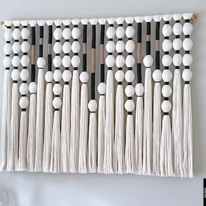 Op de afbeelding: Een grote, rechthoekige macram&eacute; wandhanger met een houten stok. Het ontwerp heeft afwisselende strengen witte kwasten, witte bollen en zwarte en bruine accenten. Het stuk is aan een lichtgekleurde muur bevestigd.
