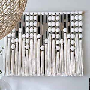 K&ouml;nnte beinhalten: Ein wei&szlig;er, schwarzer und beiger Makramee-Wandbehang mit geometrischem Muster. Der Behang ist mit wei&szlig;en Pompons und Quasten verziert.