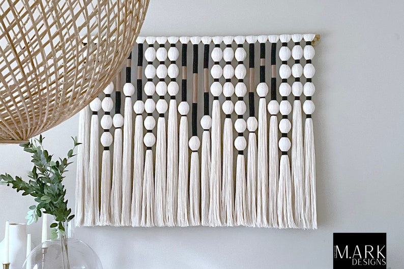 K&ouml;nnte beinhalten: Ein wei&szlig;er, schwarzer und beiger Makramee-Wandbehang mit geometrischem Muster. Der Behang ist mit wei&szlig;en Pompons und Quasten verziert.