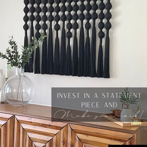 Op de afbeelding: Een zwarte macram&eacute; wandhanger met een reeks geknoopte bollen en lange kwasten. De wandhanger is opgehangen aan een houten roede met goudkleurige eindkappen. De afbeelding toont ook een houten kast en decoratieve items.