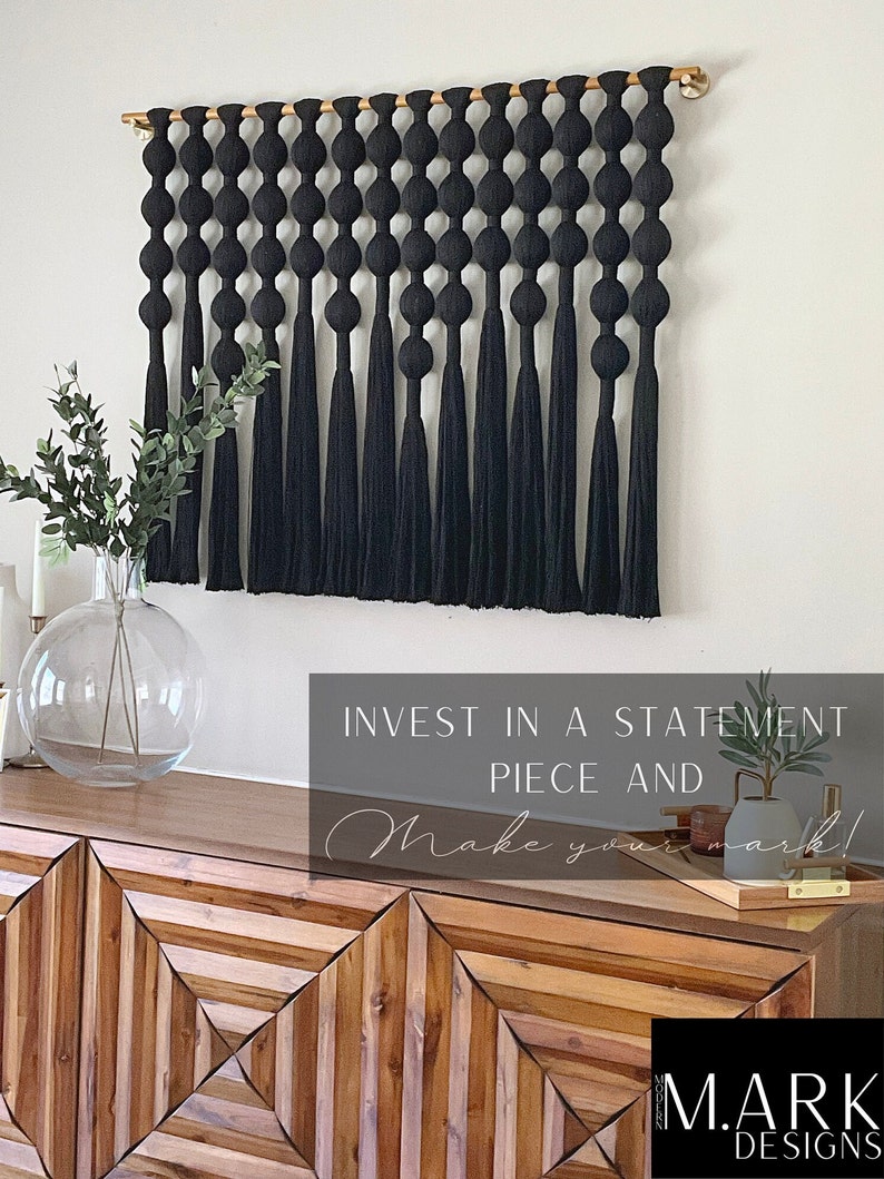 Op de afbeelding: Een zwarte macram&eacute; wandhanger met een reeks geknoopte bollen en lange kwasten. De wandhanger is opgehangen aan een houten roede met goudkleurige eindkappen. De afbeelding toont ook een houten kast en decoratieve items.