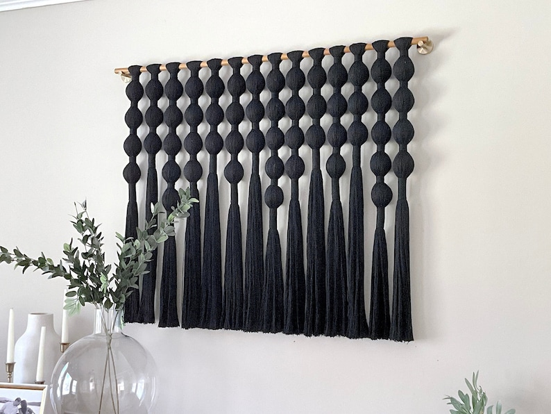 Op de afbeelding: Zwarte macram&eacute; wandhanger met een houten roede. Het ontwerp bestaat uit een reeks grote, ronde knopen die zijn verbonden door verticale strengen die eindigen in lange kwasten. Dit decoratieve stuk voegt textuur en een moderne touch toe.