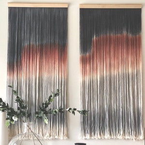 May include: Two gray, white, and orange ombre yarn wall hangings with wooden dowels at the top.  The hangings are made of long strands of yarn that are tied together.  The yarn is dyed in a gradient pattern, with the colors fading from gray to white to orange.  The hangings are hung on a white wall.