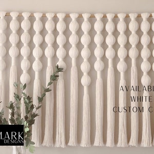 Op de afbeelding: Een grote, cr&egrave;mekleurige macram&eacute; wandhanger met een houten roede. Het ontwerp heeft een reeks verbonden bollen en lange kwasten. Tekst rechts zegt "AVAILABLE IN WHITE & CUSTOM COLORS". Een vaas met groen staat linksonder.