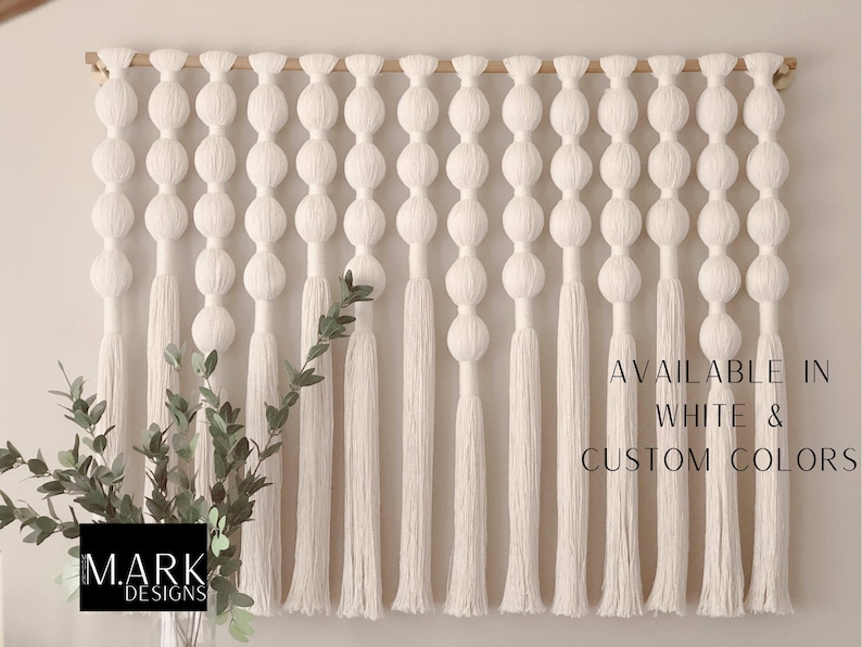 Op de afbeelding: Een grote, cr&egrave;mekleurige macram&eacute; wandhanger met een houten roede. Het ontwerp heeft een reeks verbonden bollen en lange kwasten. Tekst rechts zegt "AVAILABLE IN WHITE & CUSTOM COLORS". Een vaas met groen staat linksonder.